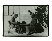 View of two men seated at a table playing checkers outside.