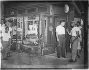 Group of men and boys standing on the sidewalk outside of a liquor store. The location is unknown, but the store is numbered 1500 and various alcohol advertisements are visible in the windows.