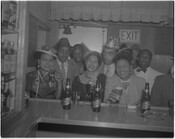 Promotional photograph for Arrow Beer in an unidentified bar. The photograph was taken from behind the bar and depicts a group of men and women holding glasses of beer.
