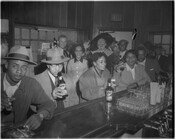 A group of patrons seated at an unidentified bar with others standing behind them, including the Hals Beer mascot, Dutch painter Franz Hals. Multiple people can be seen holding Hals Beer bottles. The image was part of a series of promotional photographs for the Baltimore, Maryland, beer company.