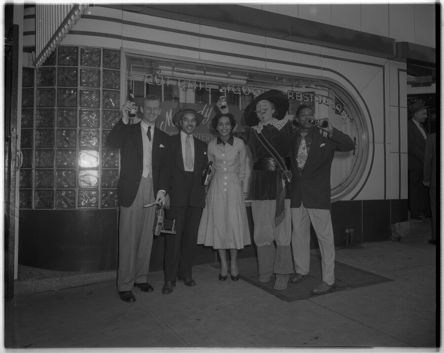 Four people posing with the Hals Beer mascot, Dutch painter Franz Hals, in front of an unidentified bar. The image was part of a series of promotional photographs for the Baltimore, Maryland, beer company.