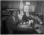 An unidentified man at a desk holding an Arrow Beer bottle with a full glass of beer in front of him.