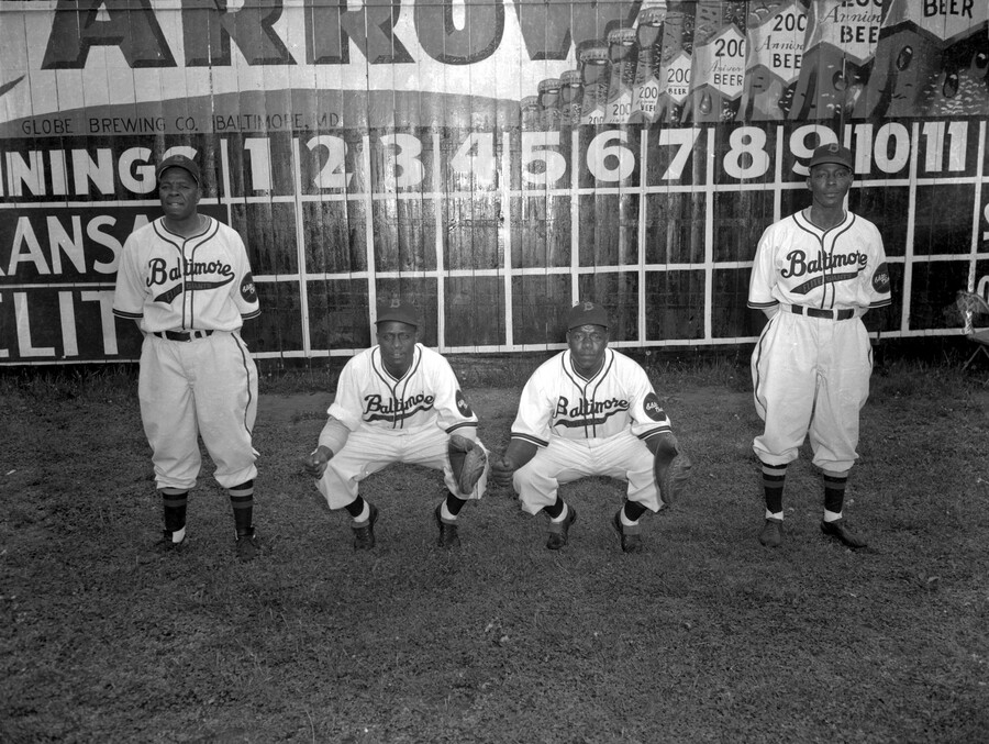Four catchers of the Baltimore Elite Giants, a Negro League baseball team, are pictured from left to right: Vic Harris (1905-1978), Hoss Walker (1904-1984), Frazier Robinson (1910-1997), and Johnny Hayes (1910-1988). The players pose in front of a scoreboard with an advertisement celebrating the 200th anniversary of Arrow Beer.