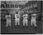 Group portrait of four batters of the Elite Giants, a Negro League baseball team from Baltimore, Maryland. Left to right: Henry Kimbo, Robert "Butch" Davis, Lester Locket, and Lenny Pearson. Behind them is a scoreboard and advertisement celebrating the 200th anniversary of Arrow Beer.