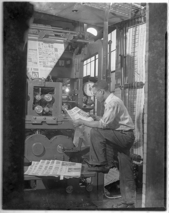 View of an unidentified man reading a freshly printed newspaper in front of a newspaper printing press.