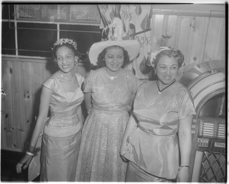 Three unidentified women in dresses and hats standing by a jukebox.
