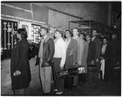 Group of unidentified men and women at work, standing in line to punch timecards. Some of the individuals are holding lunch pails.