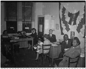 A group of unidentified men, women, and one child inside what is possibly a campaign office. The office features an image of Franklin Delano Roosevelt on the wall.