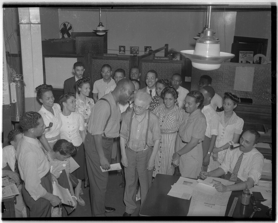 Group of men and women in an office, likely at the Afro-American newspaper building in Baltimore, Maryland. Office doors contain nameplates, "Mr. H.H. Murphy" and "Mr. B.A. Murphy."