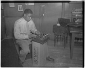 An unidentified man working on a typewriter that has been set upon a cardboard lamp box.