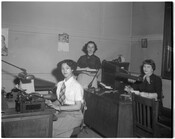 Three employees working in an office, two seated and one standing. The individual in the foreground is seated at a typewriter.