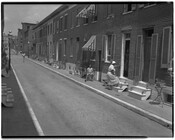 Street view of houses on an unidentified alley with residents sitting on stoops out front.