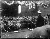 View of a large crowd seated at an unidentified outdoor event. Attendants wear formal clothing and are seated behind stanchions while a police man stands before them. Decorations above the crowd indicate the events is a celebration or festival.