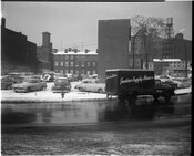 View of the parking lot at South Paca Street and Camden Street in Baltimore, Maryland with row houses in the background. The snow-covered lot is filled with cars and a Janitors Supply Houses truck passes by on the street.