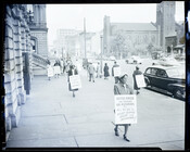 Men and women wearing sandwich boards protest on the sidewalk outside of City Hall in Baltimore, Maryland, demonstrating against a plan to turn McCulloh Street into a one-way thoroughfare. One woman holds a sign that reads, “Mister Mayor, you increase our assessments but all we get is heavy traffic noise and accidents!” An automobile is…