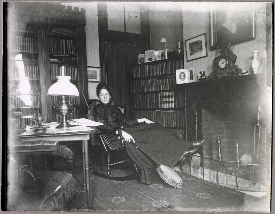 Ellen L. Duvall Steuart in her home at 123 West Lanvale Street, Baltimore, Maryland. Mrs. Steuart leans back in a rocking chair beside a fireplace, holding a banjo in her right hand.