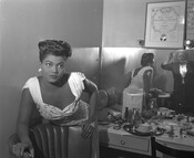 Actress and singer Pearl Bailey leaning over an armchair in her dressing room while wearing an ornate white dress. An unidentified person can be seen in the mirror behind her.