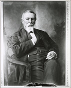 Portrait of John H. B. Latrobe seated on a wooden chair and photographed inside a Masonic Temple. Latrobe's legs are crossed and his right hand is placed inside his waistcoat.