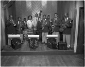 Doug's Blue Notes Band standing on a stage, including Doug McArthur, Ruby Glover, and seven other unidentified members. The male band members are posing with various instruments while Ruby Glover holds a microphone. The words "Doug's Blue Notes" and "DBN" are on geometric-shaped sheet music stands in front of the musicians.