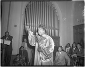 Mahalia Jackson (1911-1972) performing at a church. A microphone stand is in her left hand and her right arm is raised. The location is possibly Gillis Memorial Church in Baltimore, Maryland, as Reverend Thomas Jackson of that church stands in the background. Men and women are seated behind Ms. Jackson and a pipe organ is…