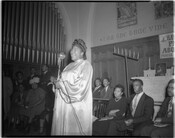 Mahalia Jackson performing at a church, possibly Gillis Memorial Church in Baltimore, Maryland, as Gillis's pastor Reverend Theodore Jackson appears in a related photograph. Men and women are seated behind Mahalia Jackson and a pipe organ is visible in the background.