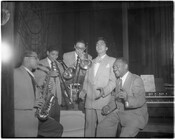 A group of unidentified musicians performing or practicing inside a club. Group includes a saxophonist, two trombonists, and two vocalists. A piano is visible in the background.