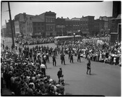 Street scene of a marching band performing during a parade, possibly on Memorial Day. A large crowd can be seen on the sidewalks, and a bus is visible in the background. The image appears to have been taken at the intersection of Broadway and Bank Streets in Baltimore, Maryland.
