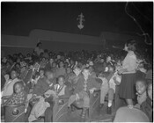 Children sitting in rows inside of a theater. Two adults are standing in the aisles watching the children. Several children in the foreground are smiling at the camera.