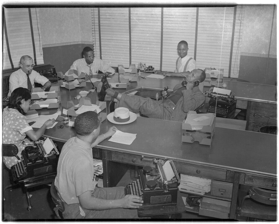 View of what was possibly the City Room of the Afro-American newspaper. Five staff members with papers and typewriters sit around a curved desk while one staffer sits with feet up, talking on the phone and smoking.