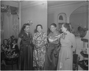 Verda Freeman Welcome, far left, standing with and talking to a group of unidentified individuals all wearing dresses. They appear to be in a living room with two other individuals seated in the background.