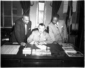 Baltimore Mayor Thomas D'Alesandro, Jr sitting at a desk and signing a document. Dr. Bernard Harris stands at left and an unidentified man stands to the right, both looking down at the document.A Baltimore surgeon who was chief of staff at Provident Hospital, Dr. Harris served on the Baltimore City Park Board and then the…