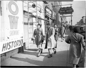 Pedestrians walking on the sidewalk of Pennsylvania Avenue in Baltimore, Maryland. A sign for Herbert's Mens Wear is hanging in the background.