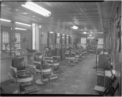 Interior view of an empty, unidentified barbershop. A row of barber chairs is set up on the left alongside a wall of mirrors.