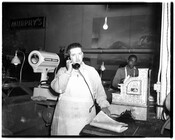 Two Murphy's grocery store employees at work. One employee is standing behind a cash register and holding a telephone to his ear. Another employee is standing in the background.