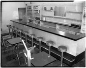 View of an empty lunch counter. There are nine stools at the counter and a row of tables to the left.