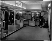 Interior of a women's clothing store. To the right, an employee is sitting on the floor measuring a customer's dress. To the left are several closets with mirrored doors.