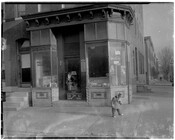 Exterior view of Phillip's Groceries located at 1161 North Calhoun Street in West Baltimore, Maryland. There are several product advertisements in the windows and a child standing on the sidewalk in front of the building. The store was owned by Philip Goldstein and was in operation at this location from 1940 to 1961.