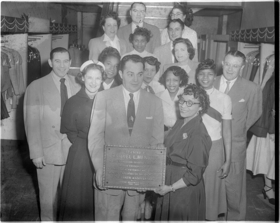 Samuel L. Hendin of Hendin's Hollywood Fashions clothing store in Baltimore, Maryland, receiving a plaque from a group of unidentified men and women. The plaque reads: "Presented to Samuel L. Hendin as an expression of esteem, affection and in recognition of outstanding achievement and leadership from the employees on the occasion of the tenth anniversary.…