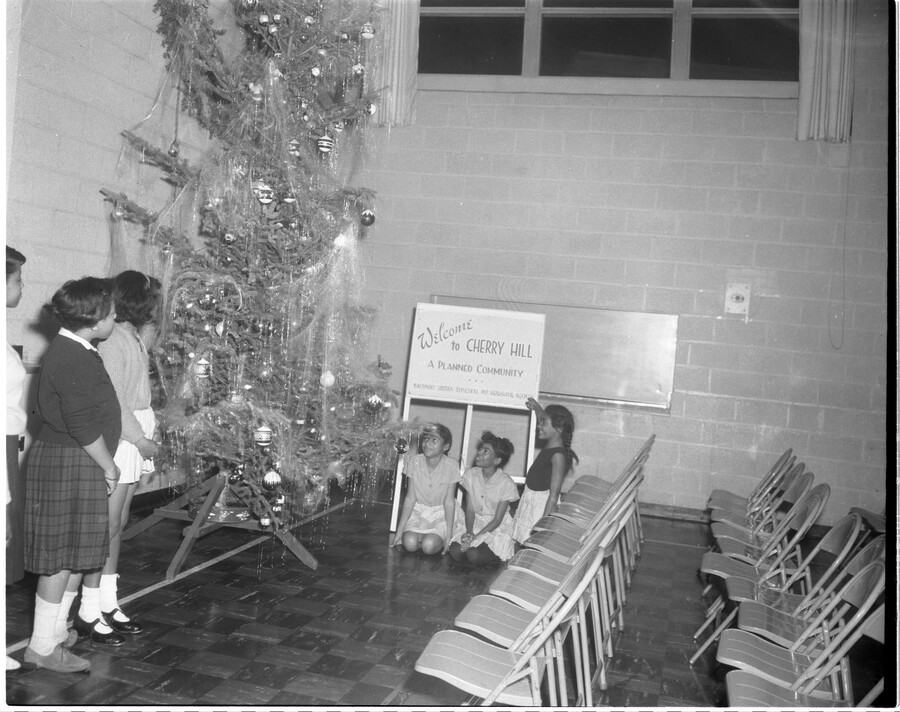Children and young adults standing and sitting around a Christmas tree with a sign that reads, "Welcome to Cherry Hill, A Planned Community."
