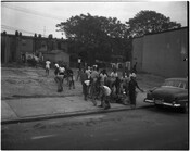 A group of children carry boxes in an open lot behind rowhomes, likely in Baltimore City, Maryland. A 1940s-era car is parked beside them.