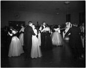 A group of young men and women dance in formal attire, possibly at a school event.