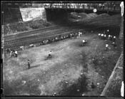 View looking down at a group of men playing a baseball game besides railroad tracks. Other men spectate from the sidelines and from a bridge above.