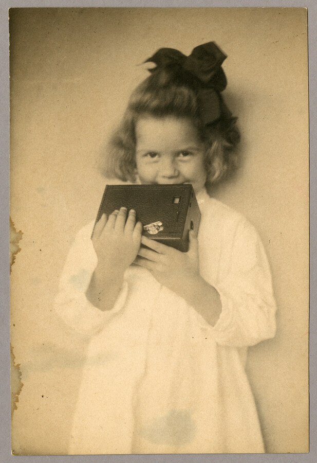 Portrait of the Baltimore, Maryland, photographer Emily Spencer Hayden's daughter Anna Bradford Hayden, also known as "Nan," holding a Brownie camera up to her face and smiling. Verso transcription: Santa Clause's best gift. Nan Hayden