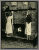 Baltimore, Maryland, photographer Emily Spencer Hayden's daughter Nan hanging stockings on the mantle of the living room fireplace with her friend Dot. Verso transcription: Nan + Dot at living room fireplace