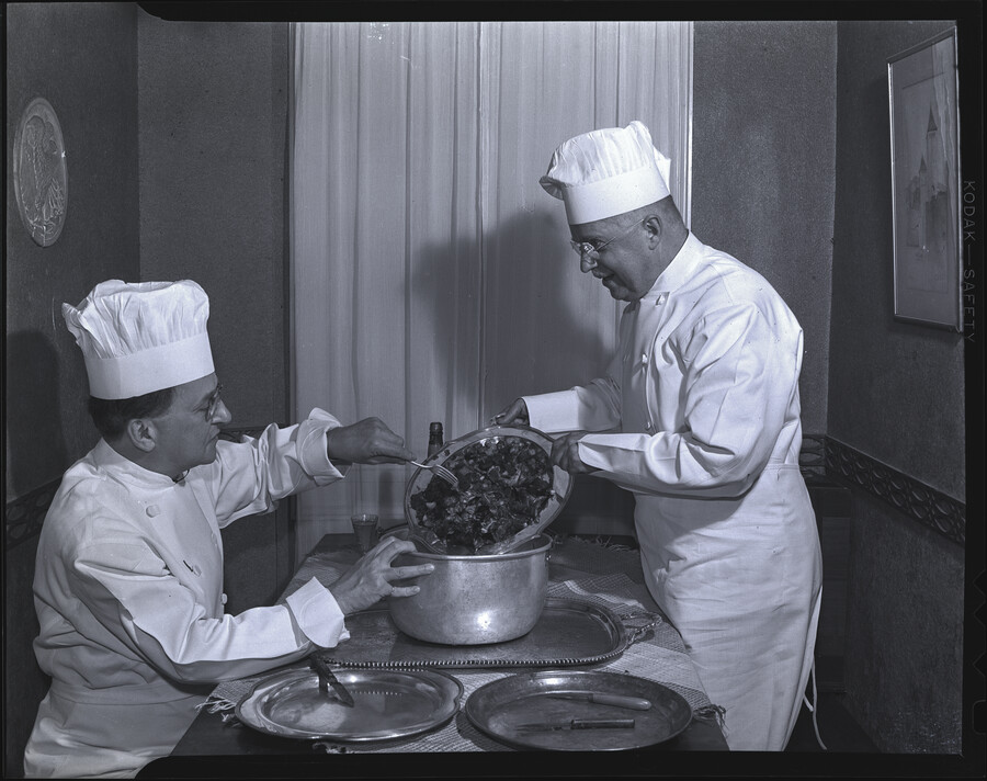 Henry P. Hopkins making a diamondback terrapin soup. Hopkins and an unidentified man are wearing chef hats and spooning ingredients into a large pot.