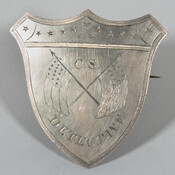 Metal badge with "C.S. Detective", crossed flags, stars, on a shield shaped badge with shirt pin on the backside, ca. 1861-1865. The flag is a variation of the national flag of the Confederate States of America (1861-1865), which had eleven stars, representing eleven states from July through November 1861. Though a later variation included thirteen…
