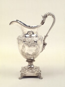 This water pitcher is decorated with a floral relief design and monogrammed with "McK" for the McKim family for whom it was made. It is further decorated with a mask head at the base of the handle and a dragon head atop the handle. The pitcher sits atop a square, claw-footed base.