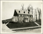 A view of the exterior of a three-story brick house located at 208 Northway in Baltimore, Maryland. The southwest corner of the Guilford Reservoir embankment is visible in the background and an automobile is parked to the left of the house.