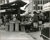 View of shoppers and stalls at Belair Market, looking southwest down Gay Street from Forrest Street in Baltimore, Maryland. The focus is on the market, but 444-448 North Gay Street buildings are visible in right background; #444 has a sign for 'Jeppi's Cafe' and #446-448 is the former Louis Kaufmann furniture store.