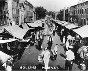 View of Hollins Market, an outdoor market at the 1000 block of Hollins Street, looking east from Arlington Street, in Baltimore, Maryland. Covered stalls line both sides of the street with vendors selling fruits, vegetables, and other goods.